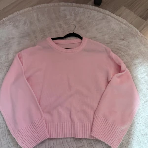 Ljusrosa stickad tröja - Säljer en ljusrosa stickad tröja med rund hals och breda ribbade muddar vid ärmslut och nederkant. Tröjan har en loose passform och är långärmad, perfekt för att ge en soft och chill vibe till din outfit.