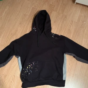Svart hoodie Lorenzo Veratti med färgstänk - Svart hoodie från Lorenzo Veratti med grå paneler på sidorna och ärmarna. Hoodien har coola färgstänk i vitt, blått, rosa och gult på axeln och fickan. Utrustad med huva och känguruficka, perfekt för en avslappnad streetstyle.