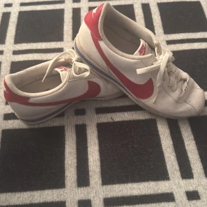 Nike Cortez vita och röda sneakers - Nike Cortez sneakers i klassisk vit färg med röda detaljer och swoosh. Låg modell med snörning och platt sula. Ovandel i skinn och syntet, rund tå och retrostil. Perfekt för dig som gillar old school vibes och sportig look.