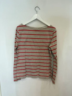 Grå långärmad randig tröja - Snygg grå långärmad tröja med röda horisontella ränder. Tröjan har en rak passform och knappar på ärmsluten som ger en extra detalj. Perfekt för dig som gillar enkel men stilren design. Materialet känns mjukt och lätt, troligen bomull eller polyester.