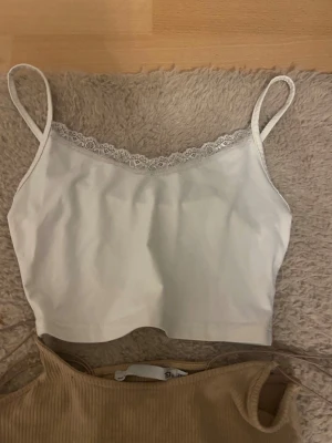 Fyra trendiga croppade toppar - Säljer fyra snygga croppade toppar: en vit spaghettitopp med spetskant, en ribbad beige linnetopp, en röd axelbandslös topp med tunna band och en vit v-ringad topp. Perfekta för sommaren och att mixa och matcha till olika outfits.