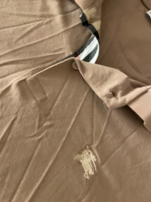 Beige Burberry pikétröja med logga - Snygg beige pikétröja från Burberry med klassisk krage och knappslå med Burberrys signaturrand på insidan. Tröjan har korta ärmar och en broderad Burberry-logga på bröstet. 