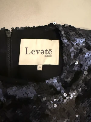 Svart paljettklänning Levete Room S - Glittrig svart kortklänning från Levete Room i storlek S. Klänningen har täckande svarta och midnattsblå paljetter över hela ytan, rund hals och korta ärmar. Perfekt för dig som vill sticka ut med en cool och festlig look. Dragkedja i ryggen för enkel påklädning.