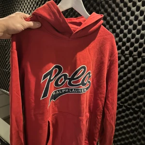 Röd hoodie från Polo Ralph Lauren - Säljer en röd hoodie från Polo Ralph Lauren med stor, broderad logga på bröstet. Tröjan har huva, lång ärm och en klassisk känguruficka framtill. Mjuk bomullskvalitet som är skön att ha på sig och passar perfekt för chill dagar.