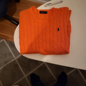 Orange stickad tröja från Polo Ralph Lauren - Snygg orange kabelstickad tröja från Polo Ralph Lauren med klassisk rund hals och svart broderad logga på bröstet. Tröjan har långärmad passform och är tillverkad i mjukt stickat material som är perfekt för kyliga dagar.