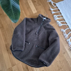 Brun teddyjacka från ARKET - Mysig brun teddyjacka från ARKET med hög krage och half zip-dragkedja. Jackan har två praktiska fickor med dragkedja framtill och är tillverkad i ett fluffigt teddy-material som håller dig varm. Perfekt för kyliga dagar och har en avslappnad passform.