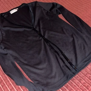 Svart kofta från Kenwel med paljetter - Svart långärmad kofta från Kenwel med v-ringning och dekorativa paljetter längs knäppningen. Snygg och enkel design som passar perfekt till jeans eller kjol. Mjukt material och smickrande passform.