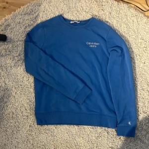 Blå sweatshirt från Calvin Klein Jeans - Snygg blå sweatshirt från Calvin Klein Jeans med vit logga på bröstet och ärmslutet. Tröjan har rund hals, ribbade muddar och är tillverkad i mjuk bomull. Perfekt för en chill och stilren look. Passar 165 - 175