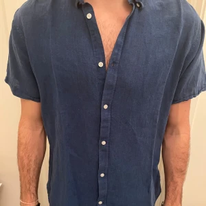 Blå kortärmad linneskjorta - Snygg blå skjorta från selected homme i mjukt linne, perfekt för varma dagar. Skjortan har korta ärmar, klassisk krage och vita knappar framtill. Enkel och stilren design som ger en chill vibe. Passar dig som gillar en avslappnad look.