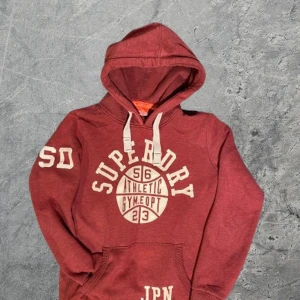 Superdry Hoodie - Säljer en röd hoodie från Superdry med stort vitt tryck framtill och på ärmen, sälj i befintlig skick. Tröjan har huva med vita snören och en klassisk känguruficka. Perfekt för dig som gillar sportig och avslappnad stil. Om du undrar något är det bara att skriva🙌✅