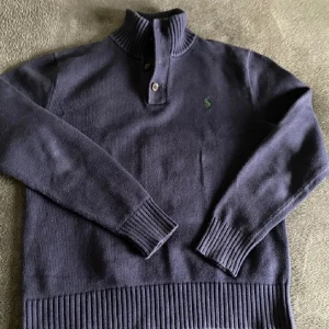 Mörkblå stickad tröja från Polo Ralph Lauren - Mörkblå stickad tröja från Polo Ralph Lauren med ribbad krage och två knappar vid halsen. Klassisk grön broderad logga på bröstet och breda ribbade muddar vid ärmslut och nederkant. Perfekt för lager-på-lager och chill stil.