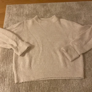 Beige ribbad stickad tröja - Mysig beige stickad tröja med ribbad struktur och högre rund hals. Tröjan har långärmade ärmar och en loose passform som ger en chill vibe. Perfekt att slänga på sig när det är kallt ute.