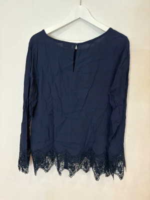 Mörkblå blus med spets från VILA - Mörkblå blus från VILA med långa ärmar och rund halsringning. Nederkanten och ärmsluten har snygga spetsdetaljer som ger en extra touch. Blusen är lätt och luftig, perfekt för dig som gillar en stilren look med lite edge.