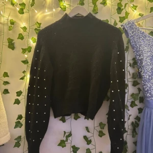 Svart stickad tröja med pärlor - Snygg svart stickad tröja med pärldetaljer längs ärmarna och hög hals. Tröjan har en croppad passform och långa ärmar, perfekt för dig som gillar att sticka ut med coola detaljer. Passar till många olika stilar och är superenkel att matcha.
