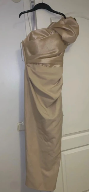 Champagnefärgad långklänning med volymärm - Snygg långklänning i champagnefärgad satin med en axelbandslös design och en stor volymärm på ena axeln. Klänningen har draperade detaljer och en elegant, figurnära passform. Perfekt för dig som vill sticka ut med en unik och lyxig look.