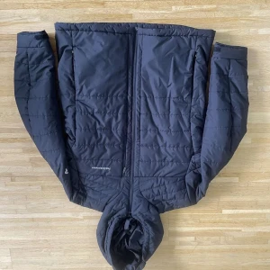 Svart pufferjacka från Peak Performance - Säljer en svart pufferjacka från Peak Performance med huva och dragkedja framtill. Jackan har quiltad design och är tillverkad i syntetmaterial som håller dig varm under kyliga dagar. Perfekt för dig som gillar stilren och funktionell ytterkläder.