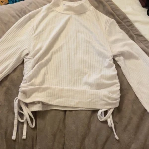 Vit ribbad polotopp med snörning - Säljer en vit ribbad polotopp med långa ärmar och snygga snörningsdetaljer på båda sidorna. Toppen har en hög krage, perfekt för att styla med jeans eller kjol. Materialet känns mjukt och bekvämt mot huden. Pris kan diskuteras och köparen står för frakt (den är liten i storleken)