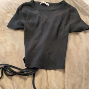 Svart ribbad topp med snörning - Svart ribbad topp med korta ärmar och snörningsdetalj i ryggen  som ger en snygg siluett. Toppen är croppad och har en rund halsringning. Perfekt att styla med höga jeans eller kjol för en trendig look. Pris kan diskuteras och köparen står för frakt (den är liten i storleken 