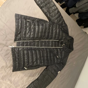 Svart Moncler dunjacka med dragkedja - Svart dunjacka från Moncler med quiltad design och vita dragkedjor. Jackan har lång ärm, ståkrage med tryckknappar och flera praktiska fickor med dragkedja. Klassisk Moncler-logga på ärmen. Perfekt för kalla dagar och snygg till streetwear. Har ej kvitto men har bokningsbekräftelse. Storlek L men kan passa som M!
