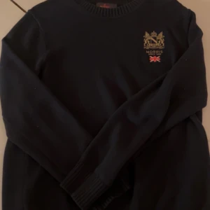 Mörkblå stickad tröja Morris L - Snygg mörkblå stickad tröja från Morris i storlek L. Tröjan har rund hals, ribbade muddar och ett broderat emblem med engelsk flagga på bröstet. Perfekt för dig som gillar klassisk stil med en twist.