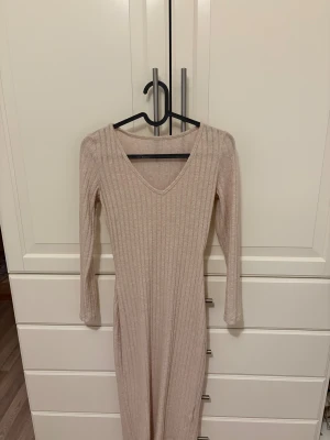 Beige ribbad långklänning med v-ringning - Säljer en beige långklänning med ribbad struktur och v-ringad hals. Klänningen har långa ärmar och är figurnära i modellen, perfekt för dig som gillar stilrena och enkla plagg med snygg passform.
