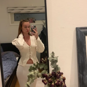 Krämvit v-ringad blus med knappar - Säljer en krämvit blus med v-ringning och långa puffärmar. Toppen har snygga knappar framtill och en lätt croppad passform med slits nedtill. Materialet känns mjukt och stretchigt, perfekt att styla med både jeans och kjol. Fråga gärna om fler bilder eller mått!