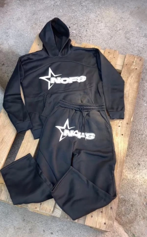 Svarta mjukisbyxor från NOFS - Svarta mjukisbyxor från NOFS med vit logga och stjärndetalj på vänster lår. Byxorna har elastisk midja med snörning för skön passform och är gjorda i mjukt syntetmaterial. Perfekta för chill eller träning, matcha med hoodie för hel look.