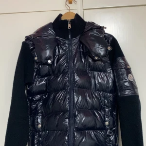 Svart pufferjacka från Moncler - Svart pufferjacka från Moncler med glansig framsida, stickade ärmar och huva. Jackan har dragkedja framtill, quiltade detaljer och Moncler-logga på ärmen. Perfekt för dig som vill ha en snygg och varm jacka med street vibe.