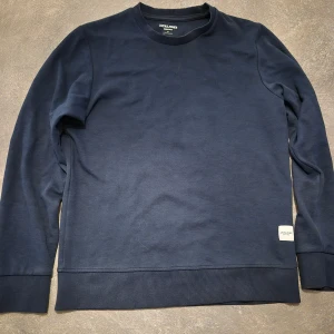 Mörkblå sweatshirt från Jack & Jones - En stilren mörkblå sweatshirt från Jack & Jones med rund hals och långa ärmar. Tröjan har en ribbad kant vid ärmslut och nederkant samt en liten vit logolapp nedtill. 