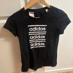 Svart Adidas t-shirt med tryck - Svart t-shirt från Adidas med vit logotyp tryckt tre gånger på framsidan. Klassisk rund hals och korta ärmar. Tillverkad i mjuk bomull som är skön att ha på sig. Perfekt för en sportig och avslappnad stil.