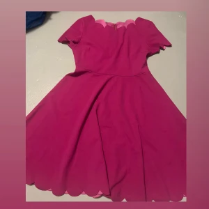 Rosa/lila kortklänning med scallop-detaljer - Klänningen har korta ärmar och en utställd kjol som ger en feminin siluett. Dragkedja i ryggen för enkel påklädning. 