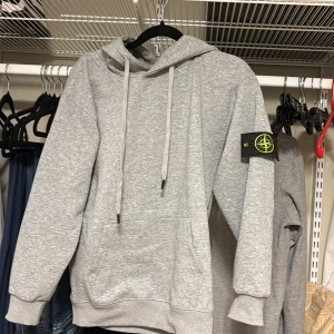 Grå hoodie från Stone Island - Grå hoodie från Stone Island med klassisk logotyp-patch på vänster ärm. Tröjan har huva med snörning, magficka och ribbade muddar. Köpt från butik för nån vecka sen men bestämde mig för att inte behålla den då den va lite kort i ärmarna💕