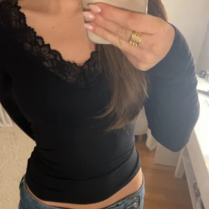 Svart långärmad topp med spets - Säljer en svart långärmad topp från Vero Moda med snygg v-ringning och spetsdetaljer runt halsen. Modellen är slim och sitter tajt mot kroppen, perfekt att styla med jeans eller kjol. Materialet är mjukt och stretchigt, troligen bomull med lite syntet.
