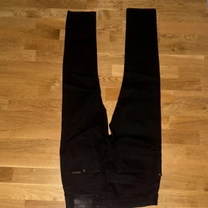Svarta jeans från Nudie Jeans - Svarta jeans från Nudie Jeans med klassisk femficksdesign och diskret logga på bakfickan. Jeansen har smal passform och raka ben, tillverkade i mjukt denimtyg. Perfekta för dig som gillar en clean och stilren look.