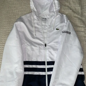 Vit och marinblå vindjacka/byxor Lacoste - Snyggt set från Lacoste i vitt och marinblått med klassiska ränder och logotyp på bröstet. Jackan har huva med snörning, hel dragkedja och sportig…kanonpris jämfört med retail priset och i nyskick