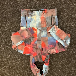 Quiksilver färgstark vindjacka med huva - Säljer en Quiksilver vindjacka med huva i regular fit. Jackan har ett unikt rutigt mönster i blått, rött, grått och lila toner. Utrustad med dragkedjor, tryckknappar och praktiska fickor. Perfekt för dig som gillar färg och vill sticka ut. Den har även ett så kallat snölås så snö inte ska komma in. Passar en som är 10-11 år