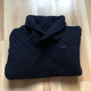 Mörkblå polotröja från Kappa - Snygg mörkblå polotröja från Kappa med diskret broderad logga på bröstet. Tröjan är stickad med strukturmönster och har långärmade ärmar samt ribbad nederkant. Perfekt för dig som gillar klassisk stil med en twist.
