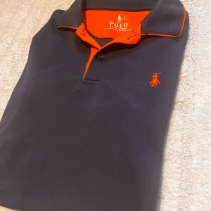 Svart och orange pikétröja Polo Ralph Lauren - Snygg svart pikétröja från Polo Ralph Lauren med orange detaljer på kragen och insidan samt orange broderad logga på bröstet. Klassisk passform med korta ärmar och mjukt bomullsmaterial. Perfekt för en stilren och sportig look. Skick 9/10. Rökfritt hus!