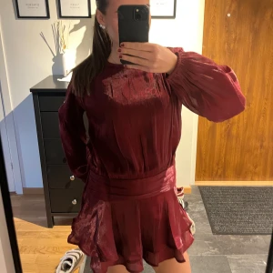 Vinröd kort klänning med volang - Vinröd kort klänning i glansigt material med långa puffärmar och markerad midja. Kjolen har volangkant och klänningen har en lös, flowy passform. Perfekt för dig som gillar en feminin och trendig look😍