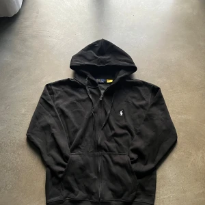 Svart hoodie från Polo Ralph Lauren - Svart hoodie med klassisk huva och snörning. Tröjan har dragkedja framtill, fickor och den ikoniska vita Polo-loggan broderad på bröstet. Tillverkad i mjuk bomull som är skön att ha på sig. Perfekt för chill dagar eller när du vill ha en clean look. Pris kan diskuteras!