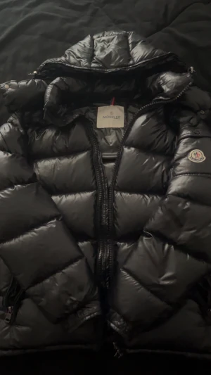 Svart Moncler pufferjacka med huva - Säljer en svart pufferjacka från Moncler med glansig finish och huva. Jackan har Moncler-logga på ärmen, dragkedja framtill och coolt serietryck på insidan. Perfekt för dig som vill ha en varm och snygg jacka med street vibe.