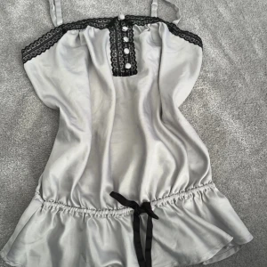 Silvrig/ljusblått linne från La Senza - Supersöt linne från La Senza med svarta spetsdetaljer och knappar framtill. Den har smala axelband, volang nertill och ett svart band i midjan. Sidenliknande material som känns lyxigt och mjukt mot huden.