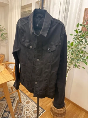 Svart jeansjacka från River Island.  - Snygg svart jeansjacka från River Island med klassisk krage och knappar framtill. Jackan har två bröstfickor med lock och långa ärmar. Perfekt att slänga på sig för en cool och enkel look.