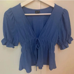 Blå blus med puffärm och knyt - Säljer en blå blus från Gina Tricot med puffiga korta ärmar och volangdetaljer vid ärmslut. Blusen har v-ringning och knyt framtill i midjan för en snygg siluett. 