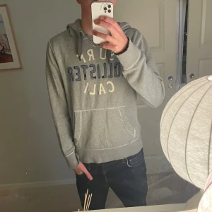 Hollister hoodie, (otroligt eftertraktad) - Grå hoodie från Hollister med broderad text 'SURF HOLLISTER CALI' i blått och vitt. Passformen är grym som ni ser på bilderna. Skicket är bra men har slitage som kommer från användning och fabrik. Sjukt eftertraktad hoodie som är ruskigt snygg! Vid fler frågor är det bara att höra av er!