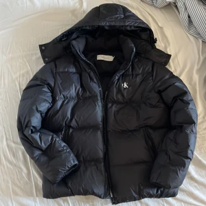 Pufferjacka Calvin Klein Jeans - Svart pufferjacka från Calvin Klein Jeans med huva och dragkedja. Jackan har CK-logga på bröstet och ett stort märke på ärmen. Den är quiltad och har två fickor med dragkedja. Perfekt för kalla dagar och har en clean, modern look. Den är som ny och har används sparsamt! Har du någons fråga så är det bara och ställa👍