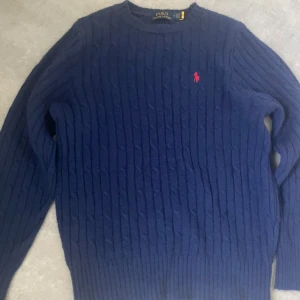 Marinblå stickad tröja Polo Ralph Lauren - Säljer en marinblå stickad tröja från Polo Ralph Lauren med klassiskt kabelstickat mönster och röd broderad logga på bröstet. Tröjan har rund hals och långa ärmar, perfekt för dig som gillar stilren och tidlös design. Storlek L men passar även mycket bra folk som har M