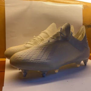 Vita fotbollsskor från Adidas - Säljer ett par fräscha vita fotbollsskor från Adidas med klassiska tre ränder på sidan. Skorna har snörning, rund tå och platt sula med metall- och plastdobbar för extra grepp på planen. Ovandelen är tillverkad i syntetmaterial med ventilerande mesh för skön känsla.