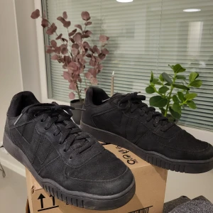 Svarta sneakers i mocka, storlek 44 - Säljer ett par helsvarta sneakers i mocka med snörning och platt sula. Skorna har en klassisk låg profil och är helt svarta, inklusive sulan och skosnörena. Perfekta för dig som gillar en clean och stilren look.