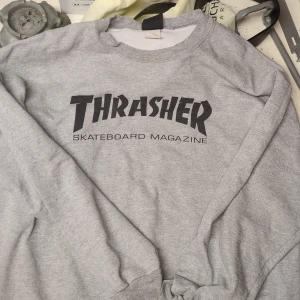 Grå Thrasher Skateboard sweatshirt  - Säljer en grå crewneck sweatshirt från Thrasher med klassisk svart logga framtill. Tröjan har rund halsringning och långa ärmar, perfekt för dig som gillar streetwear och skatekultur. Mjuk och skön bomullsblandning.